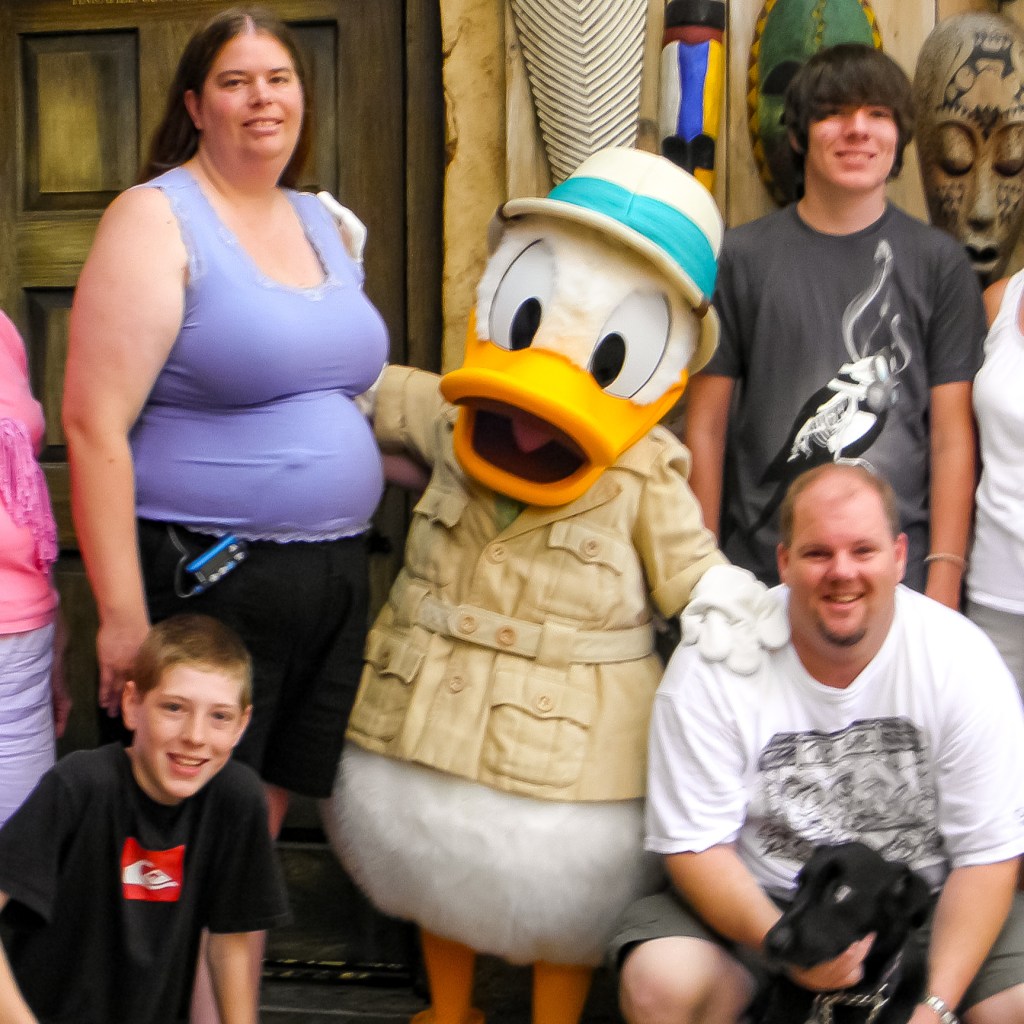 Author’s family posing with Donald Duck. Her black service dog is staring at Donald with a terrified look.